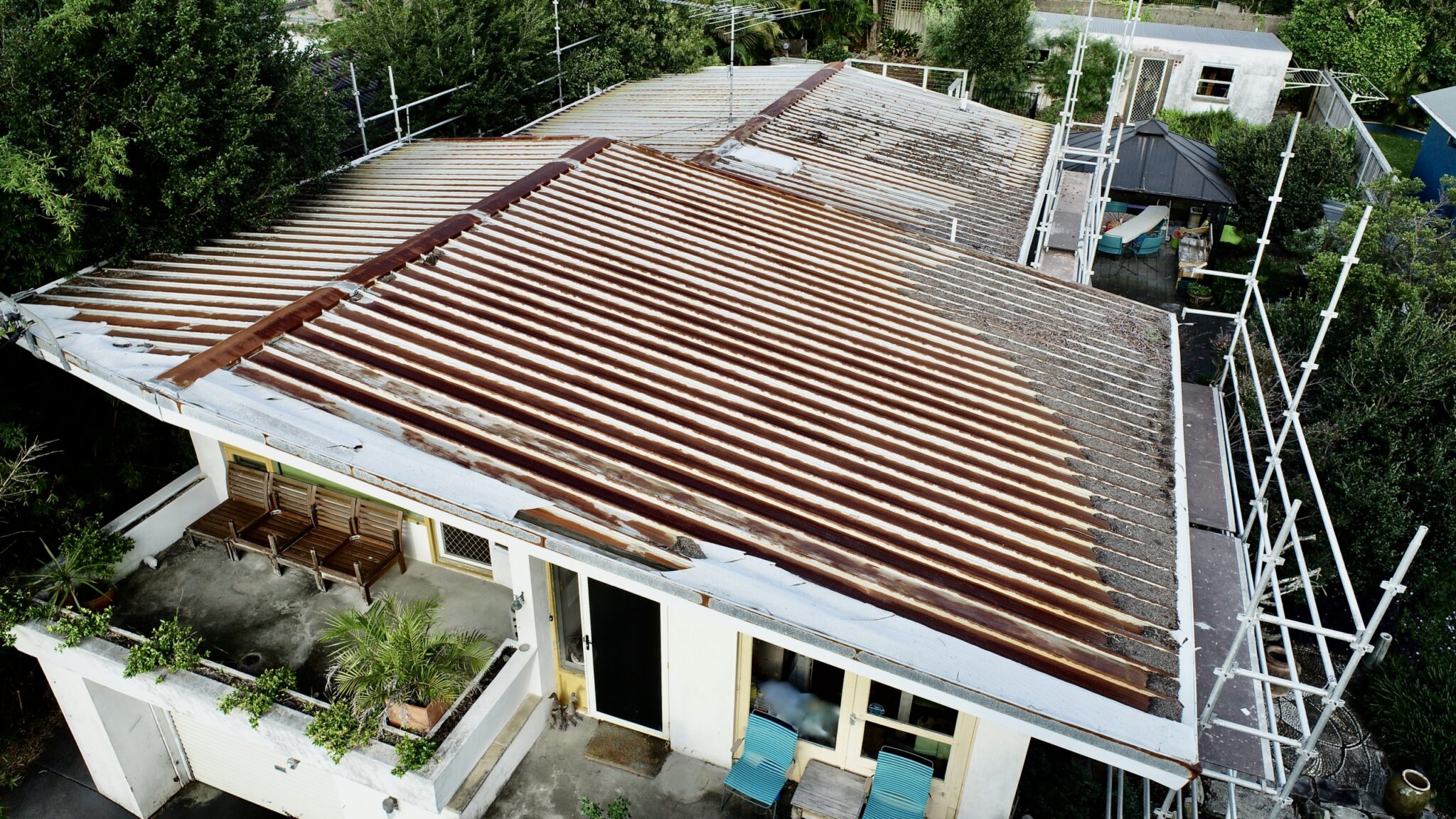 Aerial view of a house with tiles removed, exposing beams; scaffolding up for metal roofing to prevent rust.