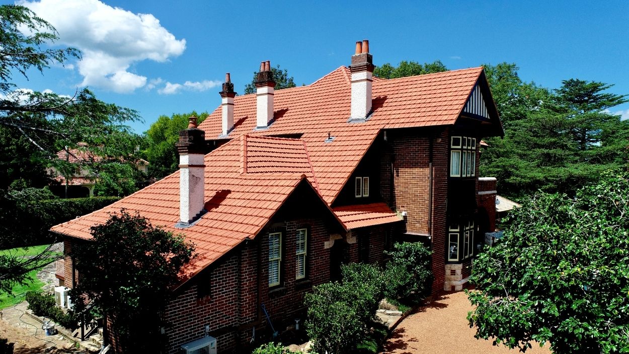 Large brick house with red tiled roof and multiple chimneys, insulated, surrounded by trees under blue sky.