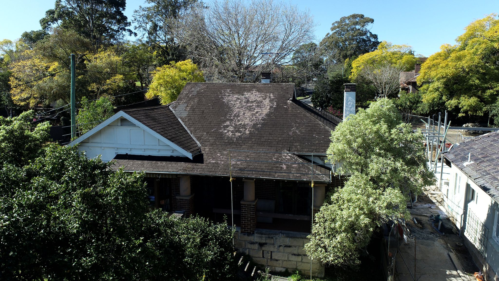 A house with a dark, patchy roof and scaffolding at its side, surrounded by trees, shows roofing before repairs finish.