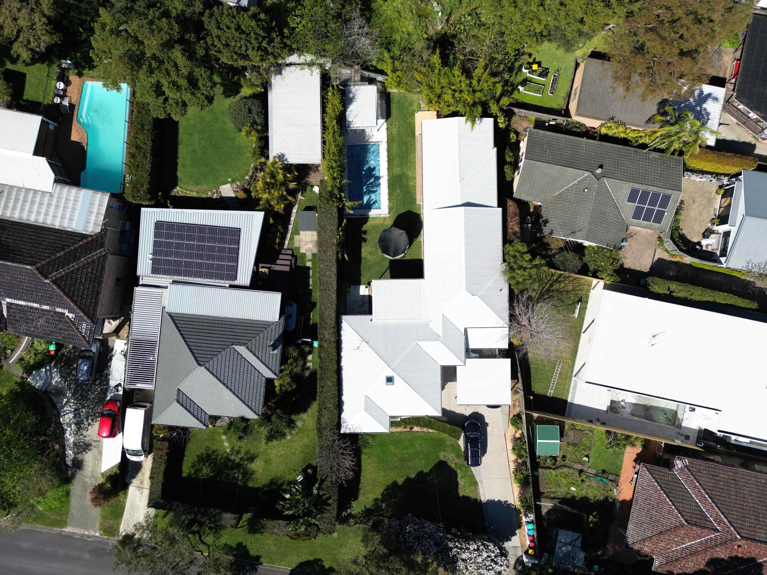 Aerial view of suburban houses with solar panels, pools, driveways, and lawns separated by fences and trees.