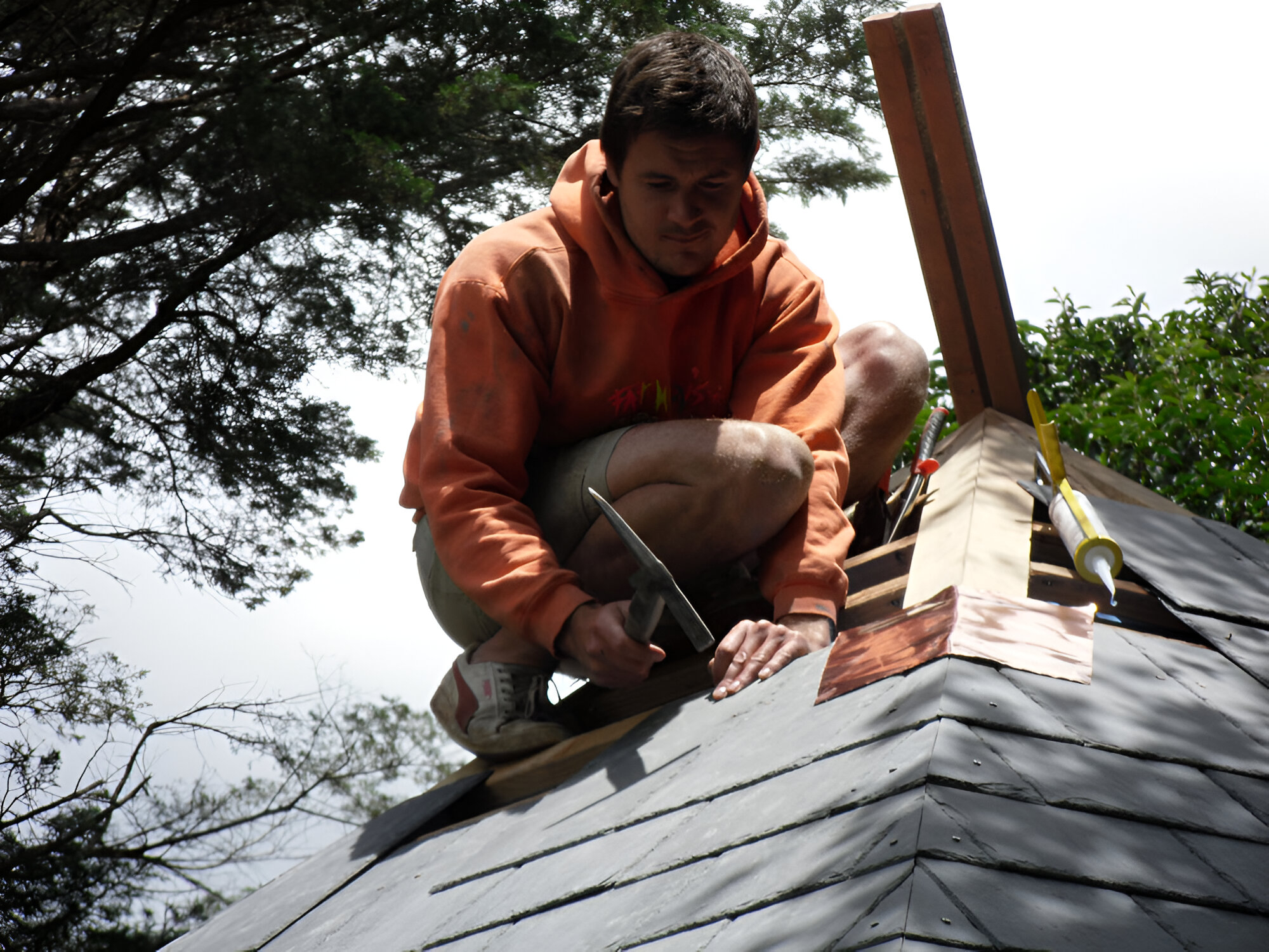 Kinsely Denning in an orange hoodie kneels on a roof, expertly hammering shingles with tools and greenery behind. 
